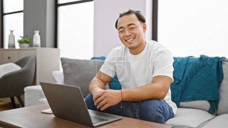 Photo for Handsome young chinese man radiates happiness, sitting on the sofa using his laptop, indulging in online technology at home in the living room area, wholeheartedly embracing the joy of indoor comfort. - Royalty Free Image