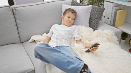 Foto de Adorable niño rubio relajándose en el sofá en casa, concentrado en ver la televisión en un cómodo estilo de vida en el interior - Imagen libre de derechos