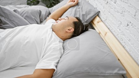 Jeune chinois épuisé en pyjama confortable trouve un confort doux dans un sommeil heureux, couché sur un lit moelleux au milieu de la lumière chaude du matin qui coule à travers l'intérieur de la chambre intime de la maison.