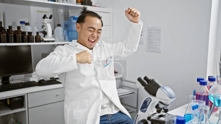 Téléchargez les photos : Jeune scientifique chinois souriant célébrant une découverte majeure au microscope en laboratoire, incarnant l'esprit gagnant de la recherche médicale - en image libre de droit