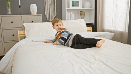 Téléchargez les photos : Jeune garçon blond souriant sur un lit blanc dans une chambre à coucher moderne intérieur - en image libre de droit