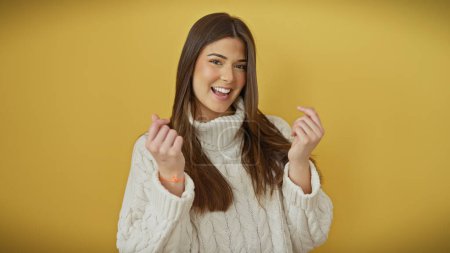 Téléchargez les photos : Une jeune femme joyeuse fait un geste d'argent sur un fond jaune vif isolé, dépeignant le style et la positivité. - en image libre de droit
