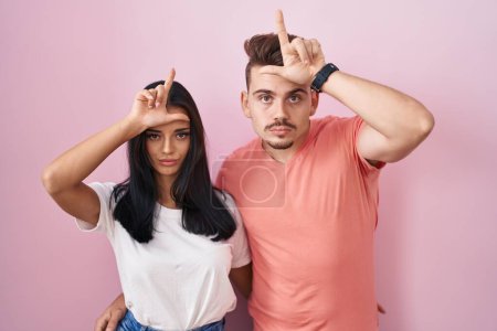 Photo for Young hispanic couple standing over pink background making fun of people with fingers on forehead doing loser gesture mocking and insulting. - Royalty Free Image