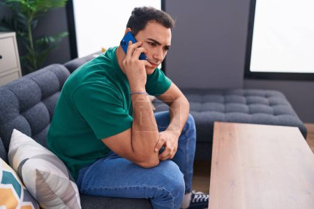 Téléchargez les photos : Jeune homme hispanique parlant sur smartphone avec une expression inquiète à la maison - en image libre de droit
