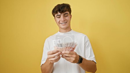 Selbstbewusster junger hispanischer Teenager, der isländische Kronen-Banknoten zählt, glücklich lächelnd vor leuchtend gelbem Hintergrund