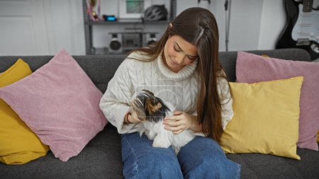 Photo for A young hispanic woman cuddles with a biewer yorkshire terrier on a couch indoors, surrounded by colorful pillows. - Royalty Free Image