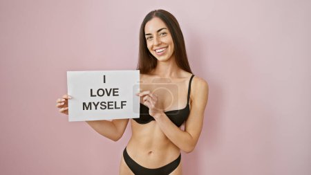 Beautiful, confident young hispanic woman in sexy lingerie stands looking proud over isolated pink background, holding a 'i love myself' banner - a stunning portrait of self-love and positivity