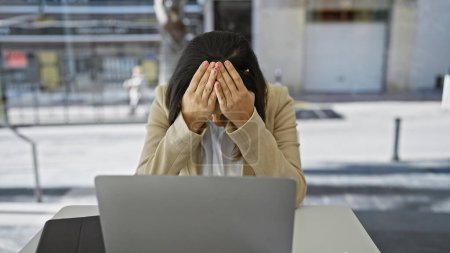 Foto de Mujer estresada sentada en una computadora al aire libre con la cabeza en las manos, mostrando frustración laboral en un entorno urbano. - Imagen libre de derechos