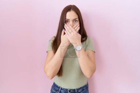 Foto de Hermosa mujer morena de pie sobre fondo rosa impactada cubriendo la boca con las manos por error. concepto secreto. - Imagen libre de derechos