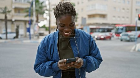 Foto de Mujer africana sonriente con trenzas usando un teléfono inteligente en una calle de la ciudad. - Imagen libre de derechos