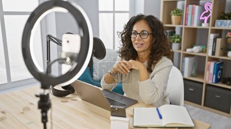 Téléchargez les photos : Femme d'âge moyen souriante enregistrant podcast dans un home studio avec un ordinateur portable, microphone, et la lumière de la bague - en image libre de droit