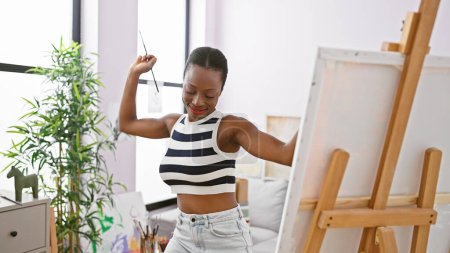 Foto de Confiada mujer afroamericana artista, bailando y sonriendo en el estudio de arte mientras disfruta y aprende pintura en una clase de creatividad - Imagen libre de derechos