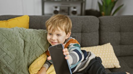 Foto de Un niño rubio alegre sosteniendo un teléfono inteligente mientras está sentado en un sofá con almohadas en una acogedora sala de estar. - Imagen libre de derechos