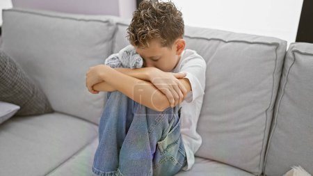 Foto de Adorable niño rubio abrazando oso de peluche, llorando solo en el sofá, expresando tristeza, estrés y desesperación en casa - Imagen libre de derechos