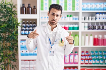 Handsome hispanic man working at pharmacy drugstore pointing down looking sad and upset, indicating direction with fingers, unhappy and depressed. 