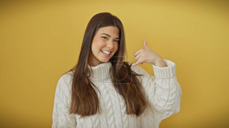 Foto de Sonriente mujer hispana joven gesto me llaman contra un fondo aislado de color amarillo brillante en un suéter blanco. - Imagen libre de derechos