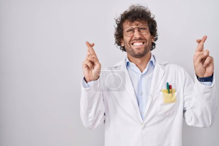 Photo for Hispanic young man wearing doctor uniform gesturing finger crossed smiling with hope and eyes closed. luck and superstitious concept. - Royalty Free Image