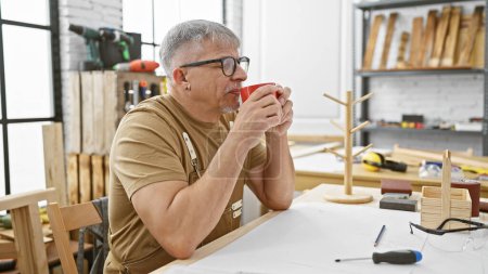 Foto de Hombre de mediana edad bebe café en un taller de carpintería, reflexionando sobre la carpintería con herramientas alrededor. - Imagen libre de derechos