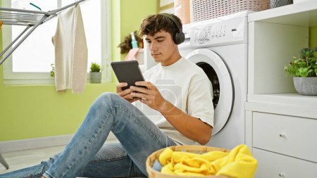 Téléchargez les photos : Cool jeune adolescent hispanique absorbé par la musique, en utilisant le pavé tactile et écouteurs en attendant la machine à laver dans la buanderie confortable - en image libre de droit