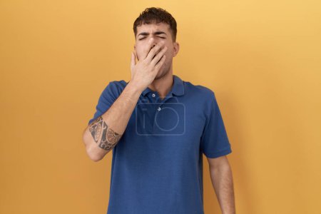 Foto de Joven hombre hispano de pie sobre fondo amarillo aburrido bostezando cansado cubriendo la boca con la mano. inquietud y somnolencia. - Imagen libre de derechos