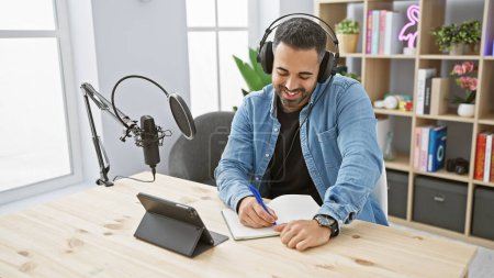 A smiling man with headphones in a podcast studio writes notes, depicting a casual indoor radio recording scene.