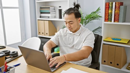 Foto de Joven hombre chino serio, un profesional de negocios enfocado, inmerso en el trabajo con su computadora portátil en el escritorio de la oficina - Imagen libre de derechos