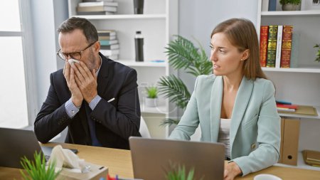 Téléchargez les photos : Deux travailleurs d'entreprise éternuent ensemble au bureau alors qu'ils travaillent dur sur un ordinateur portable, une expression claire de la grippe de bureau affliction à l'intérieur. - en image libre de droit
