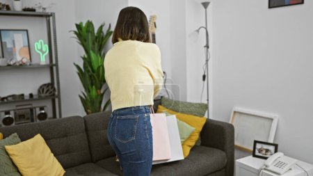 Téléchargez les photos : Une jeune femme dans un pull jaune et un jean se tient dans un salon confortable, faisant allusion à un style de vie décontracté moderne. - en image libre de droit