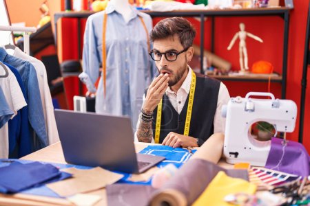 Photo for Young hispanic man with tattoos dressmaker designer using laptop at tailor room covering mouth with hand, shocked and afraid for mistake. surprised expression - Royalty Free Image