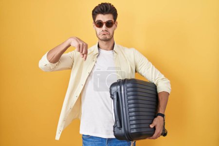 Jeune homme hispanique tenant une valise allant en vacances d'été pointant vers le bas regardant triste et contrarié, indiquant la direction avec les doigts, malheureux et déprimé. 