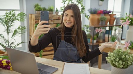 A young woman takes a selfie at a flower shop filled with plants, showcasing her business with a warm smile.