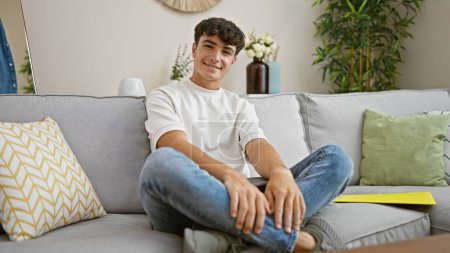 Foto de Joven adolescente hispano alegre irradia confianza mientras se relaja y sonríe en un sofá acogedor, disfrutando de la comodidad del hogar. - Imagen libre de derechos