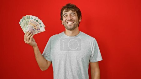Foto de Joven y sonriente hombre sosteniendo con confianza y contando su riqueza en billetes argentinos, aislado sobre un vibrante fondo rojo - Imagen libre de derechos