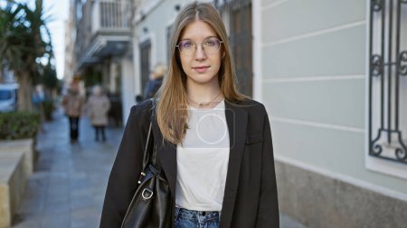 Eine junge kaukasische Frau mit Brille und blonden Haaren steht selbstbewusst auf einer städtischen Straße.