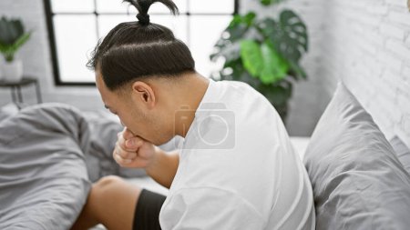 Téléchargez les photos : Jeune homme chinois assis sur le lit toussant dans la chambre - en image libre de droit