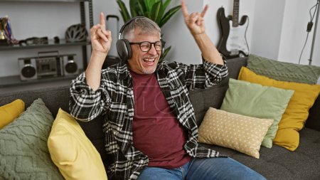 Téléchargez les photos : Un homme joyeux d'âge moyen avec les cheveux gris aime la musique sur les écouteurs dans un salon confortable - en image libre de droit