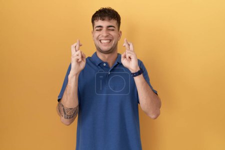 Photo for Young hispanic man standing over yellow background gesturing finger crossed smiling with hope and eyes closed. luck and superstitious concept. - Royalty Free Image