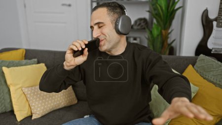 Téléchargez les photos : Homme hispanique chantant karaoké avec smartphone et écouteurs dans un salon confortable, se sentant joyeux et diverti. - en image libre de droit