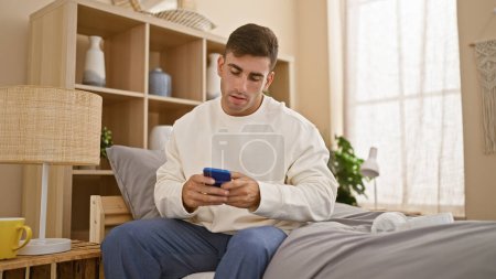 Retrato cautivador de un joven hispano, atractivo sin esfuerzo, cómodamente ubicado en su santuario de dormitorio, absorto en el universo digital, escribiendo un mensaje temprano en la mañana en su teléfono inteligente.