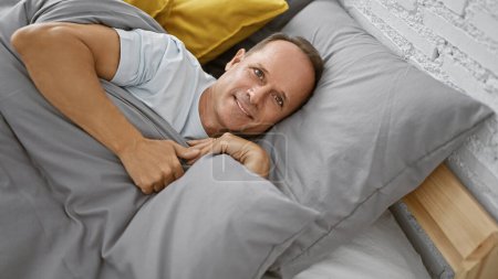 Téléchargez les photos : Beau, confiant homme d'âge moyen rayonne de bonheur à l'intérieur de sa chambre confortable, confortablement couché dans son lit, souriant le matin - en image libre de droit
