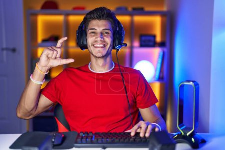 Téléchargez les photos : Jeune homme hispanique jouant à des jeux vidéo geste souriant et confiant avec la main faisant signe de petite taille avec les doigts à la recherche et la caméra. concept de mesure. - en image libre de droit