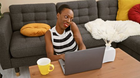 Captivating african american woman engaged in lively talk over phone, beautifully poised on home sofa, confidently juggling work on laptop while savoring morning espresso