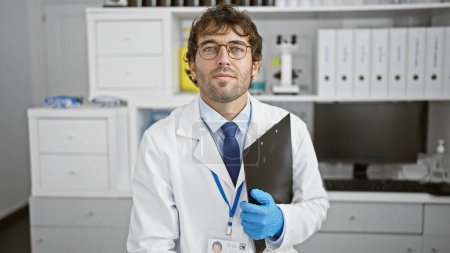 Photo for Handsome young hispanic man, focused scientist with blond beard, analyzing biology research on a clipboard while sitting relaxed at his lab table, immersed indoors in the high-tech medical laboratory. - Royalty Free Image