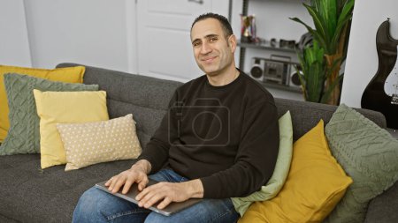Foto de Un hombre hispano relajado se sienta cómodamente en un sofá gris en una sala de estar moderna, sosteniendo una tableta. - Imagen libre de derechos