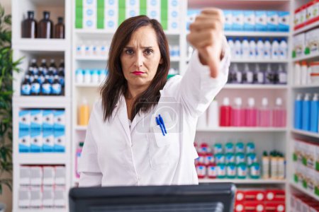 Photo for Middle age brunette woman working at pharmacy drugstore looking unhappy and angry showing rejection and negative with thumbs down gesture. bad expression. - Royalty Free Image