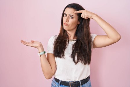 Photo for Young brunette woman standing over pink background confused and annoyed with open palm showing copy space and pointing finger to forehead. think about it. - Royalty Free Image
