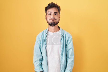 Photo for Young hispanic man with tattoos standing over yellow background puffing cheeks with funny face. mouth inflated with air, crazy expression. - Royalty Free Image