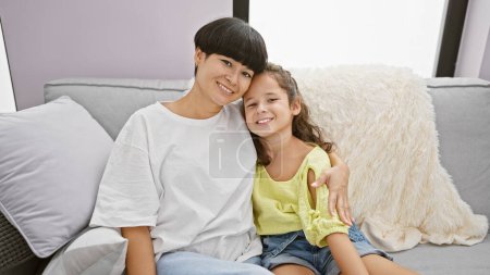 Foto de Madre e hija confiadas compartiendo un abrazo alegre, sonriendo de corazón mientras están sentadas en el sofá de la sala de estar en casa una hermosa expresión de amor familiar - Imagen libre de derechos