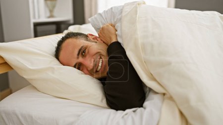 A smiling young man nestled in bed sheets expressing comfort and relaxation in a cozy bedroom setting.