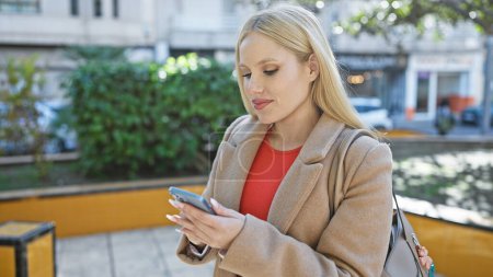 Téléchargez les photos : Une jeune belle femme blonde portant un manteau regarde son téléphone à l'extérieur dans la ville - en image libre de droit